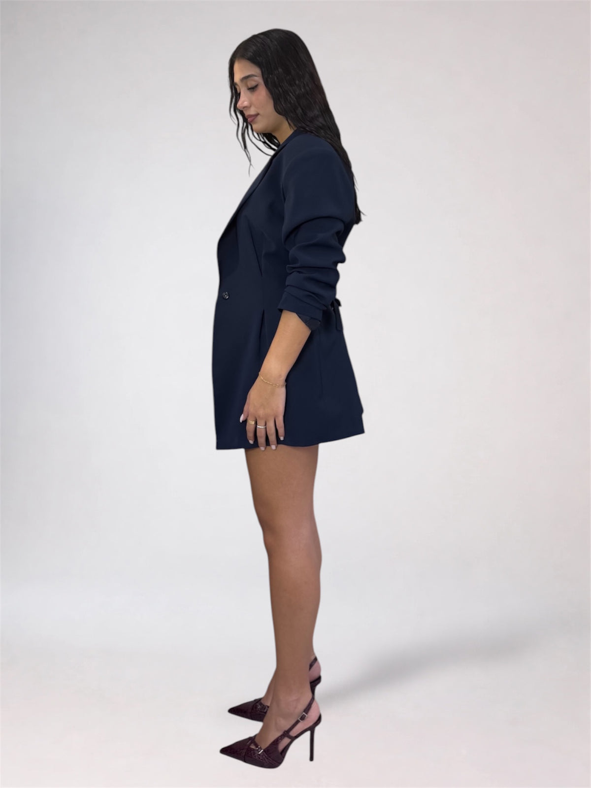 A woman stands in profile wearing a dark navy blazer-style mini dress with long sleeves and pointed heels against a plain light gray studio background.