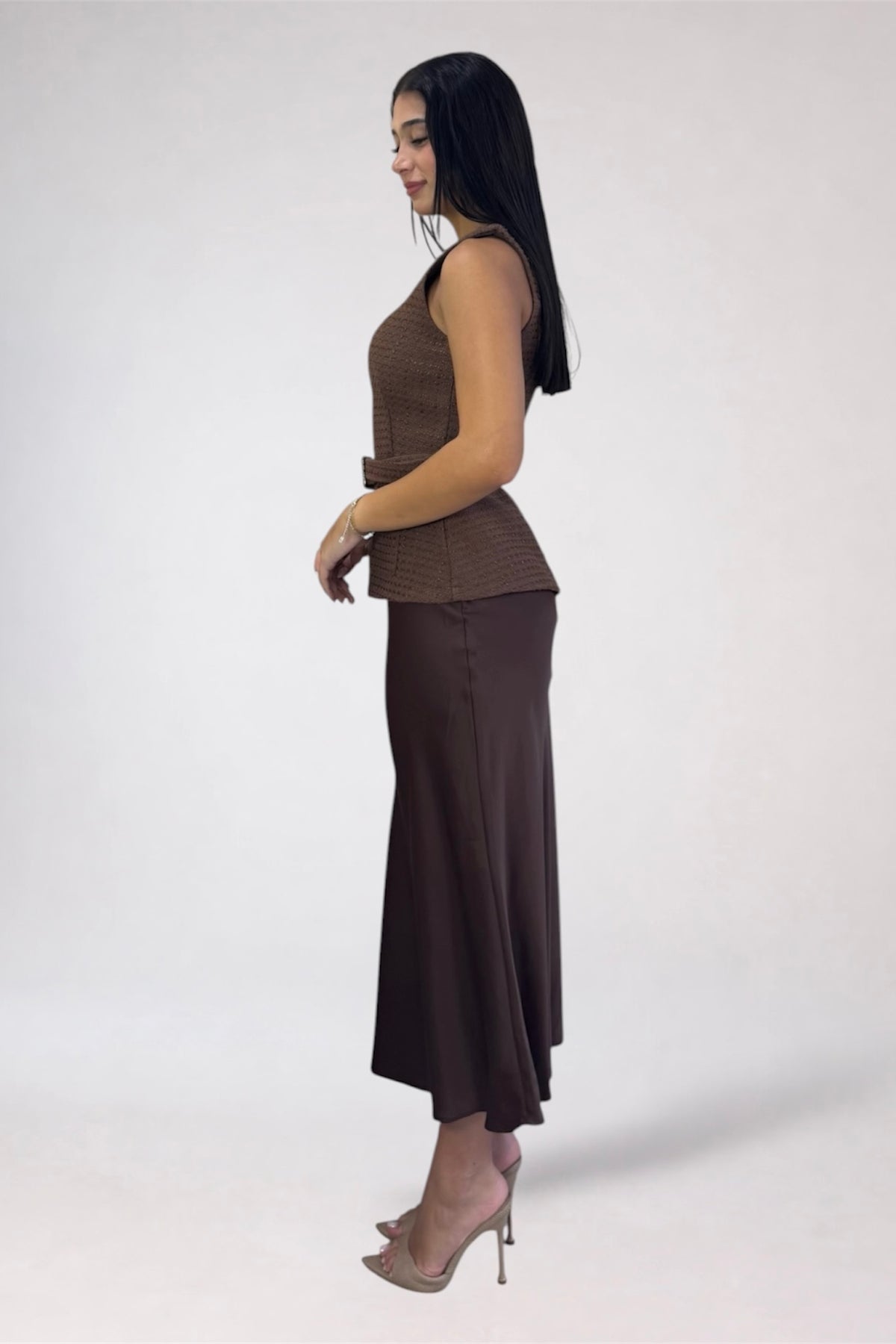A woman modeling a two-piece brown outfit consisting of a sleeveless ribbed knit top and a matching midi skirt, paired with nude high heels, against a plain light gray studio background.