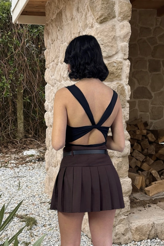 Person wearing a black top and brown pleated skirt standing in front of a stone wall.