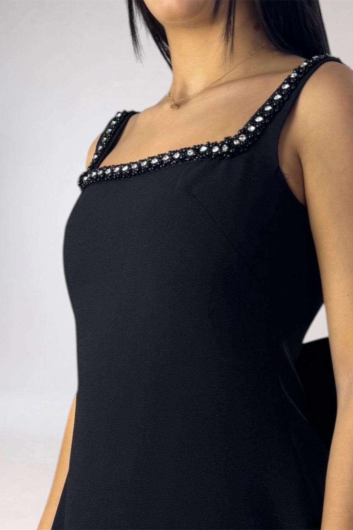 A close-up of a model wearing a black sleeveless dress with a square neckline and a decorative border of black beads and white stones along the straps, set against a plain light gray background.