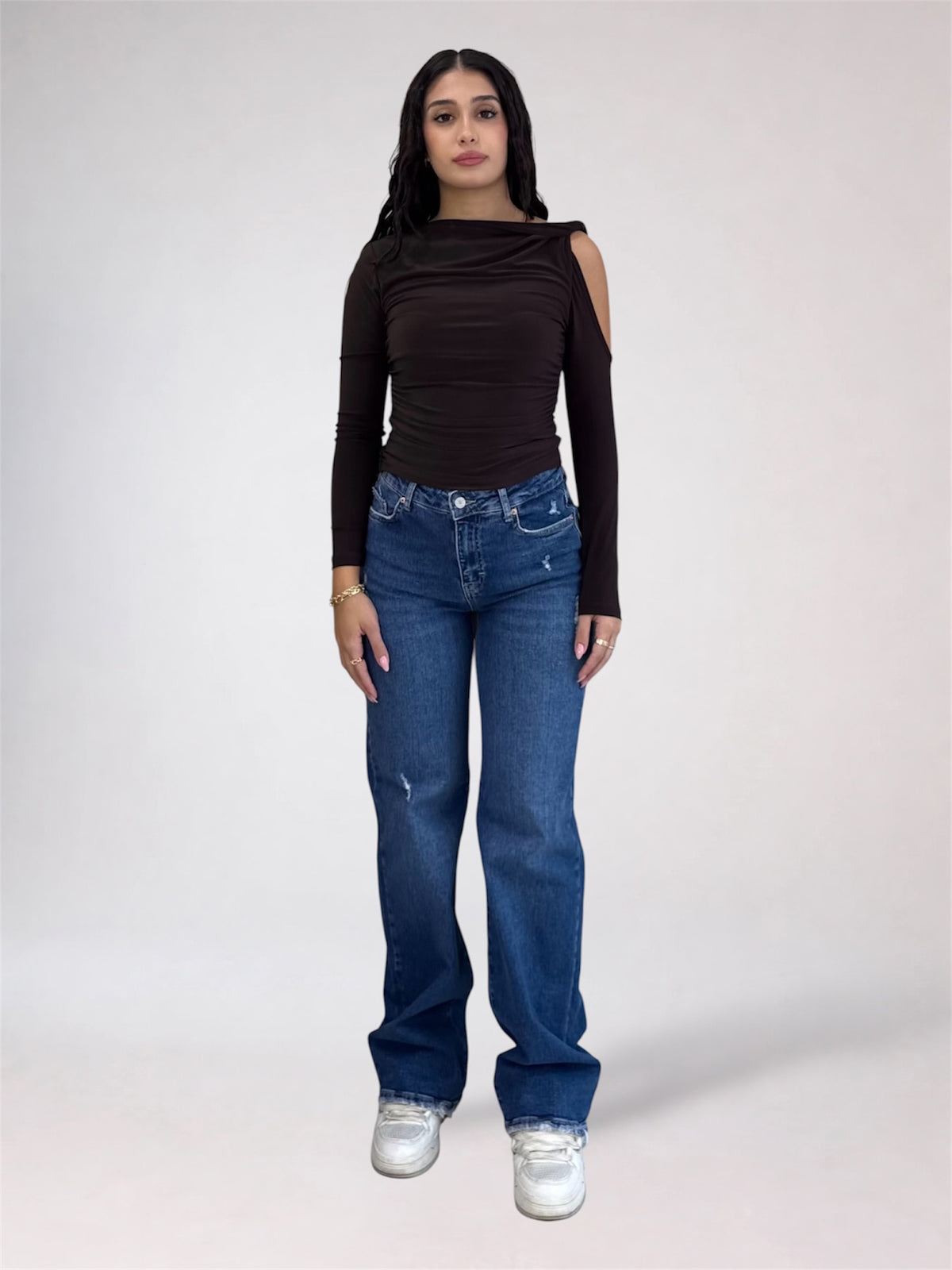 A woman modeling a dark brown long-sleeve top with shoulder cutouts and blue jeans, standing against a light gray studio background.