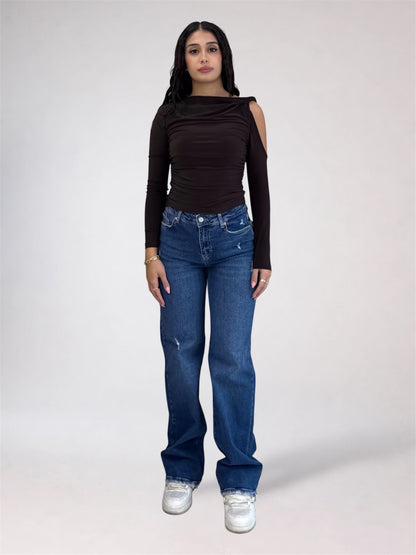 A woman modeling a dark brown long-sleeve top with shoulder cutouts and blue jeans, standing against a light gray studio background.