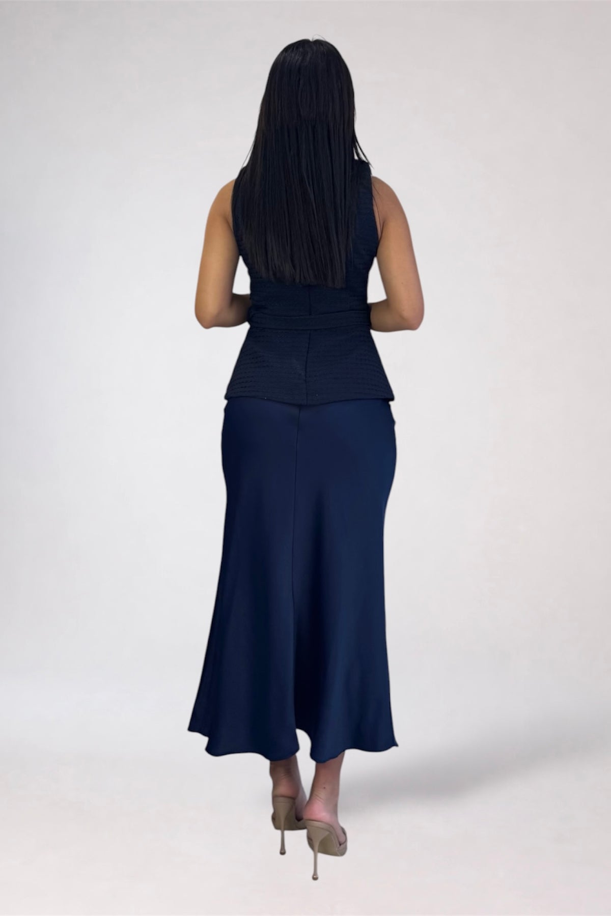 A woman viewed from behind wearing a sleeveless navy blue dress with a structured peplum waist and flared skirt, paired with beige high heels, against a plain light gray studio background.