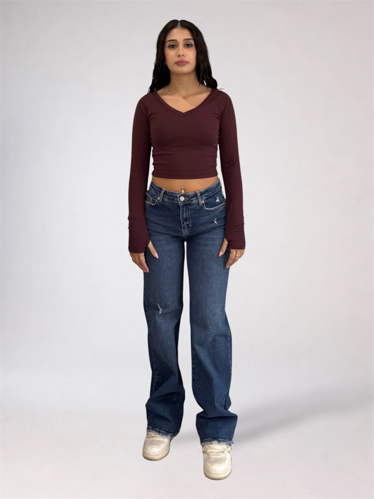 A woman modeling a long-sleeve maroon crop top and blue jeans against a plain light gray studio background.