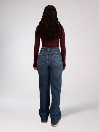 A woman viewed from behind wearing a dark burgundy long-sleeve crop top and high-waisted, wide-leg blue jeans against a plain light gray studio background.