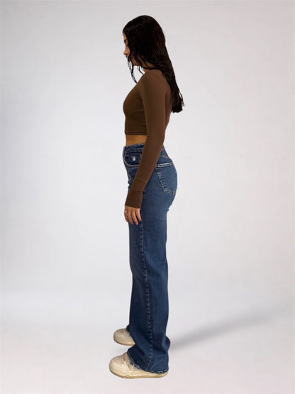 A woman viewed from behind wearing a dark brown long-sleeve crop top and high-waisted, wide-leg blue denim jeans against a plain light gray studio background.