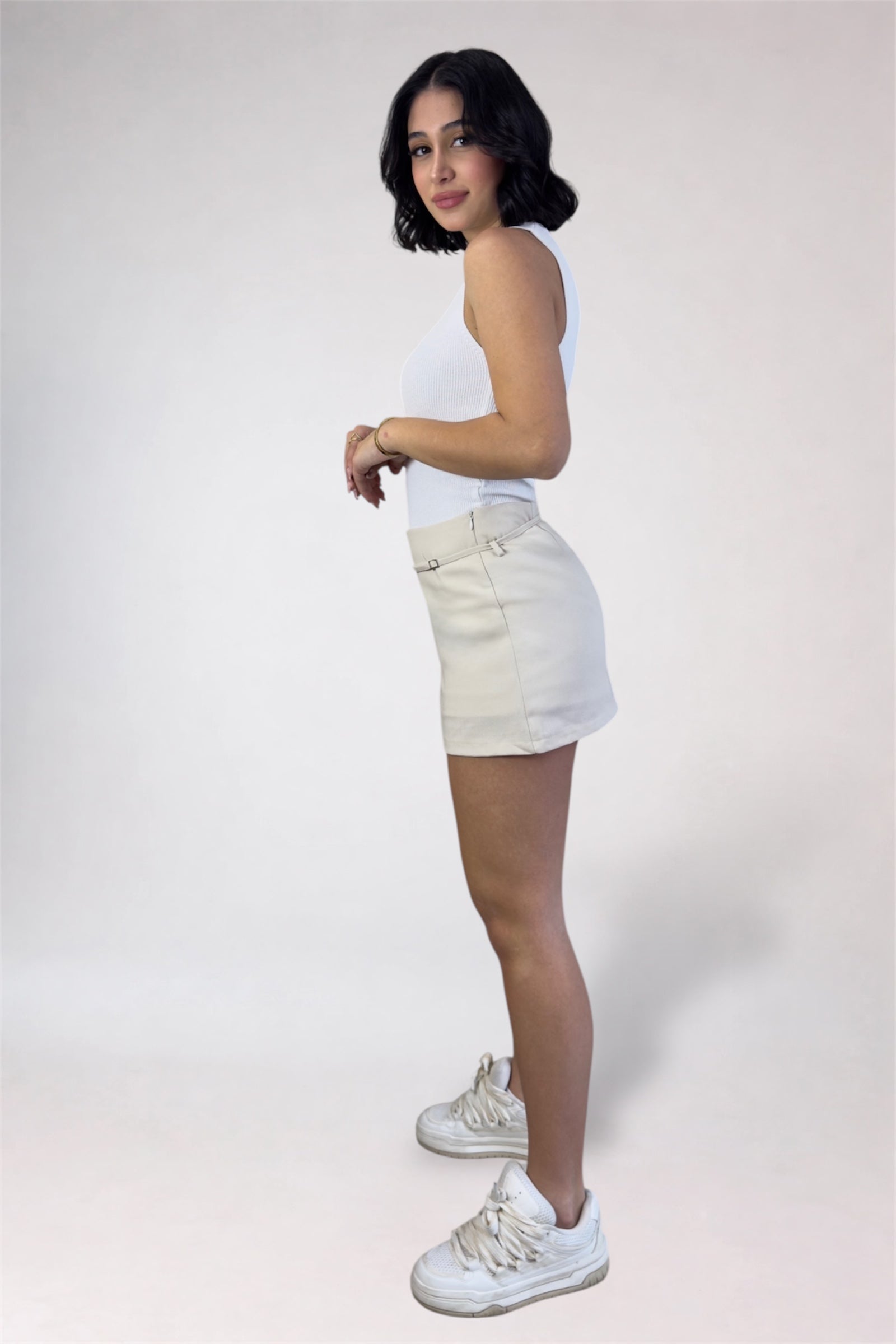 Person wearing a white sleeveless top and beige skirt on a plain background
