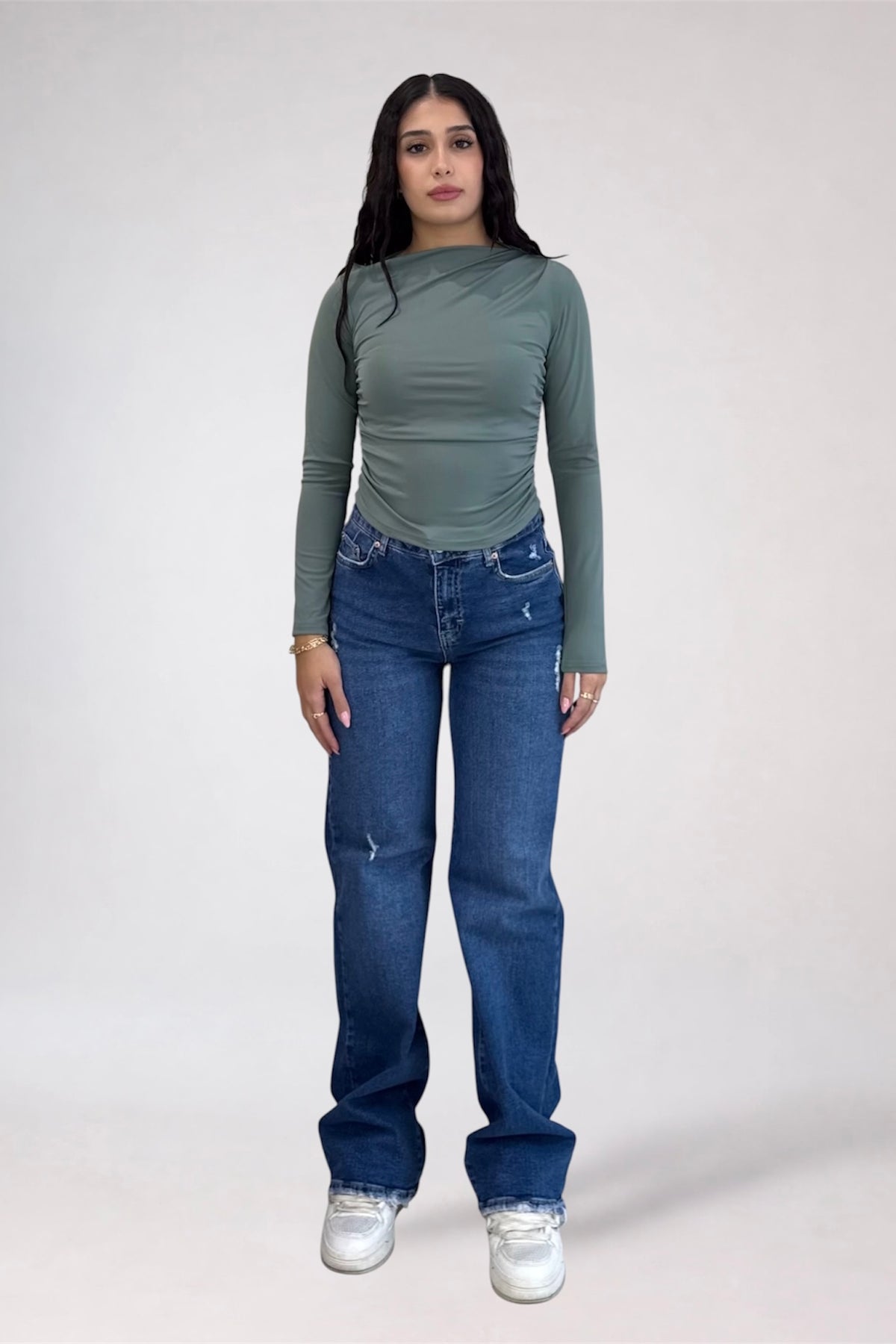 A woman modeling a sage green long-sleeve top and blue jeans against a plain light gray studio background.