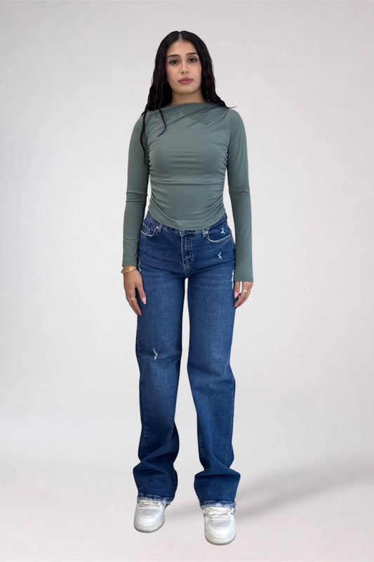 A woman modeling a sage green long-sleeve top and blue jeans against a plain light gray studio background.