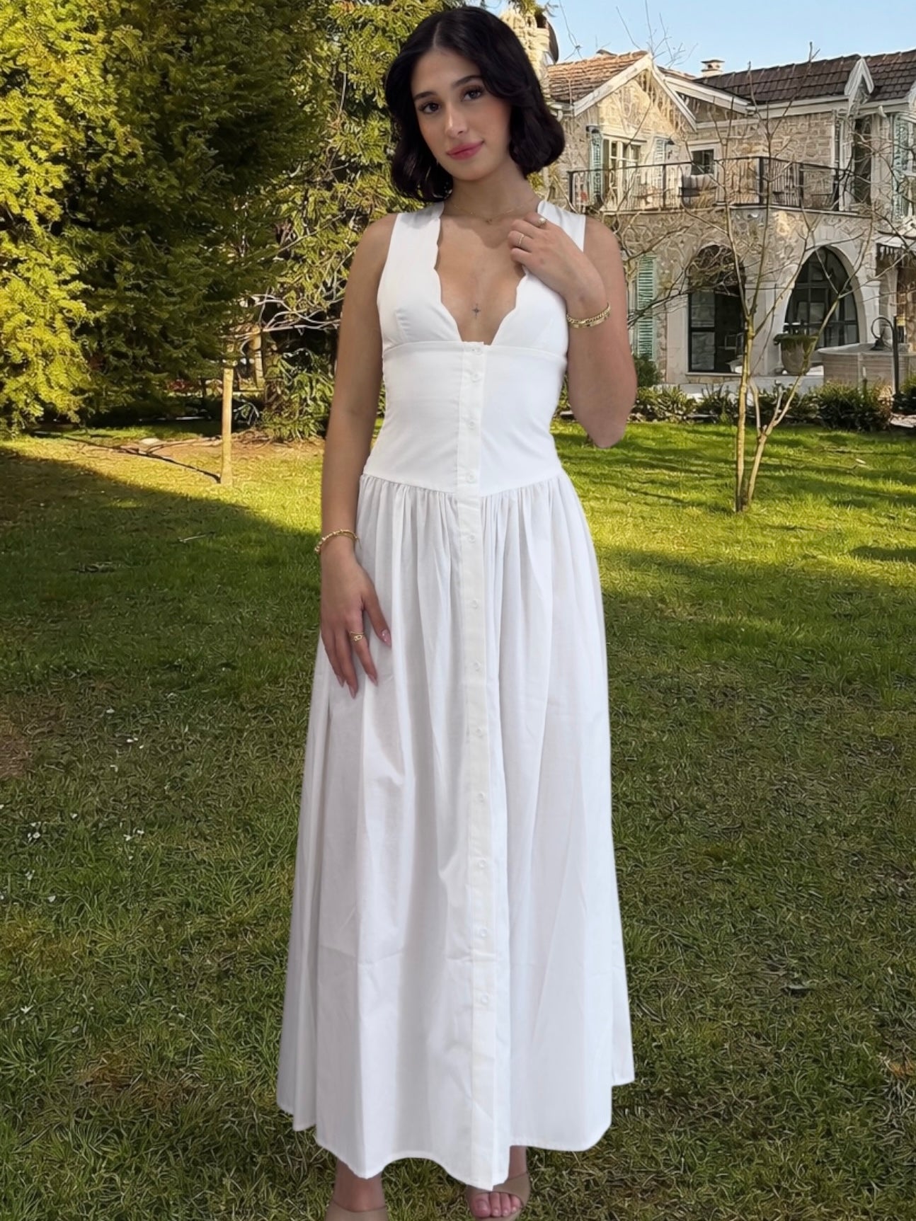 Woman in a white dress standing in a grassy area with trees and a building in the background