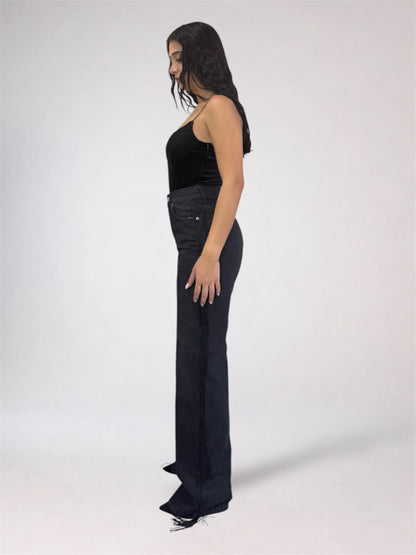 A woman stands in profile wearing a black sleeveless top and dark wide-leg jeans against a plain light gray studio background.