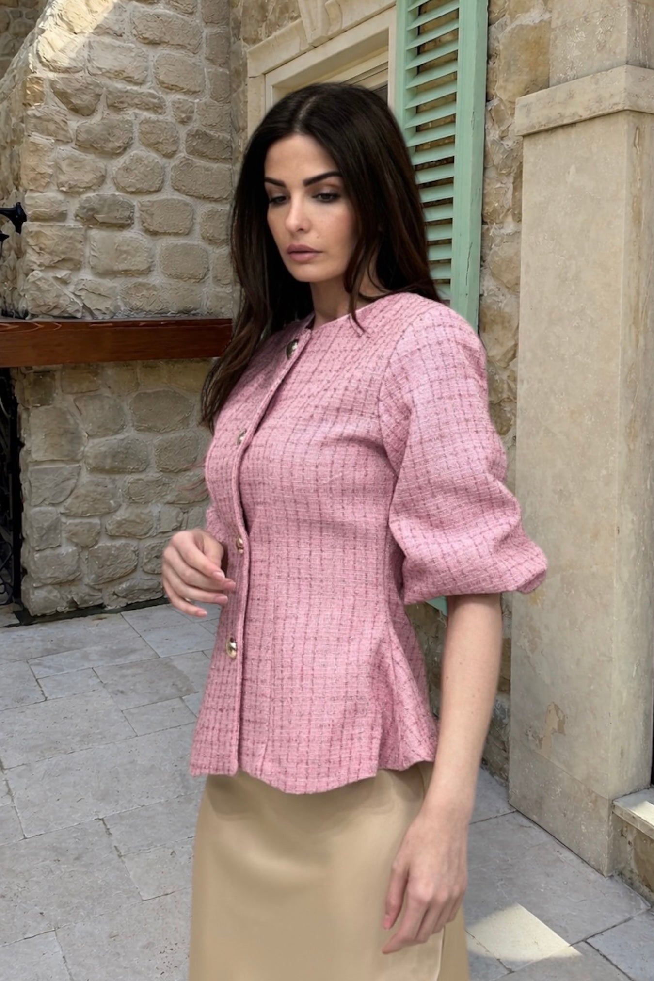 Woman wearing a pink checkered jacket and beige skirt standing in front of a stone wall.