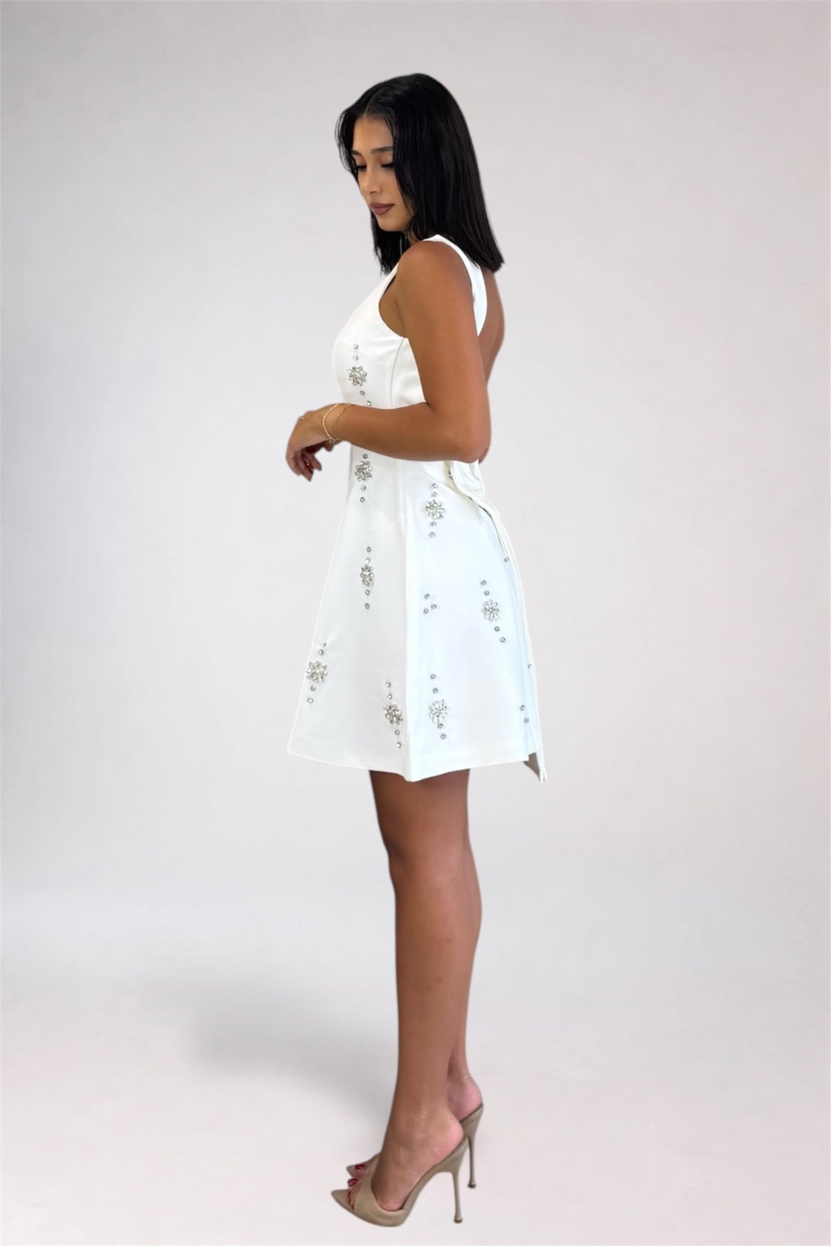 A woman modeling a sleeveless white mini dress with delicate silver floral embroidery, shown from a side profile against a light gray studio background.