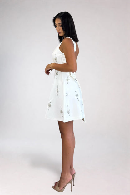 A woman modeling a sleeveless white mini dress with delicate silver floral embroidery, shown from a side profile against a light gray studio background.