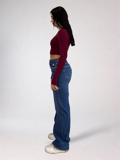 A woman viewed from behind wearing a dark burgudy long-sleeve crop top and high-waisted, wide-leg blue denim jeans against a plain light gray studio background.