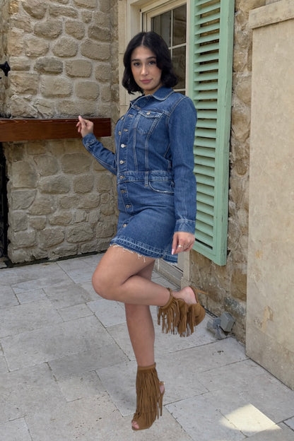 Woman in a denim dress posing in front of a stone building with green shutters.