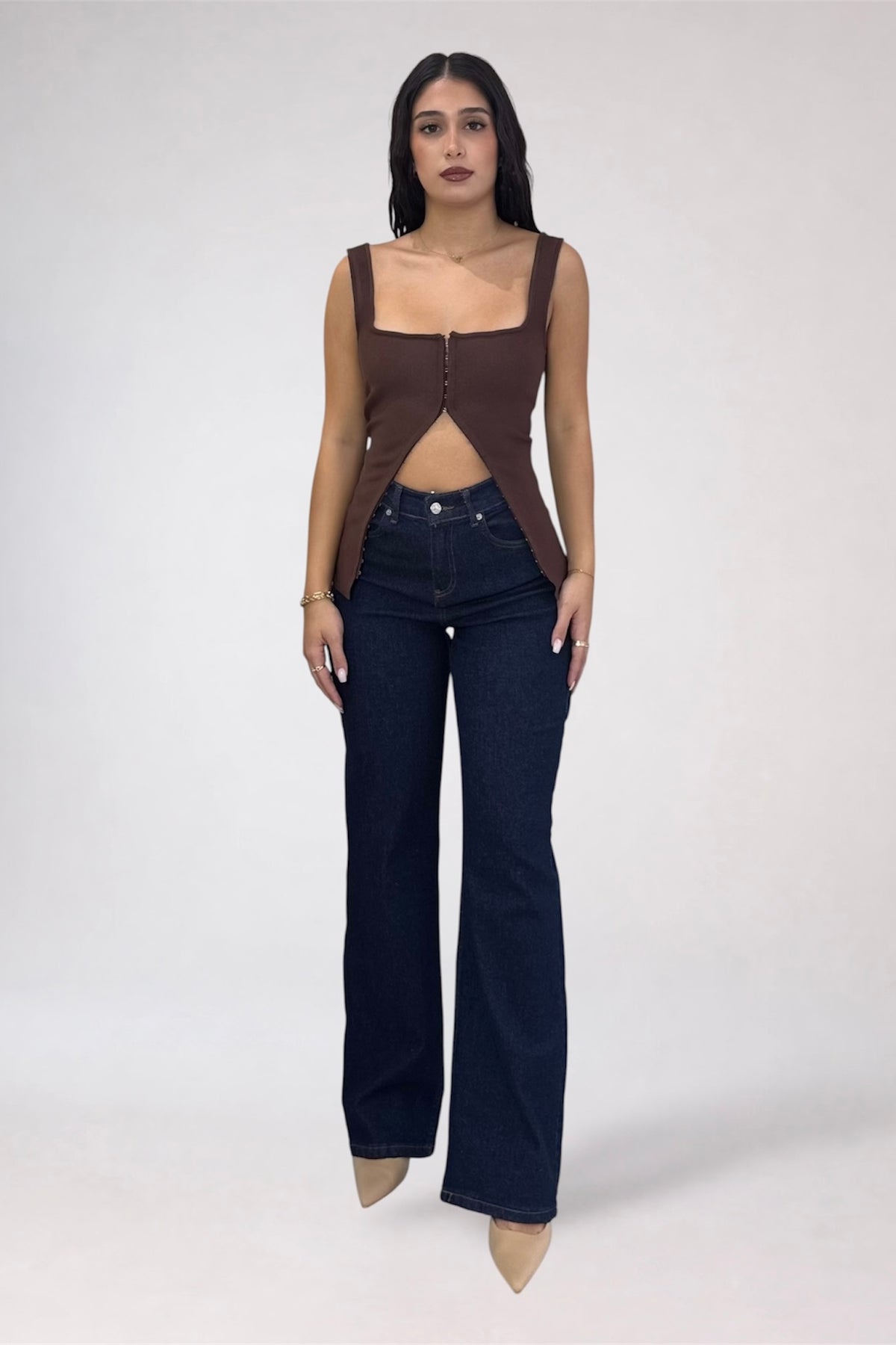 A woman modeling a brown sleeveless cutout top and dark blue flare jeans against a light gray studio background.