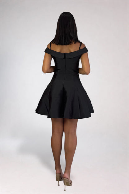 A woman viewed from behind wearing a short off-the-shoulder black dress with a flared skirt, paired with beige high heels, against a light gray studio background.