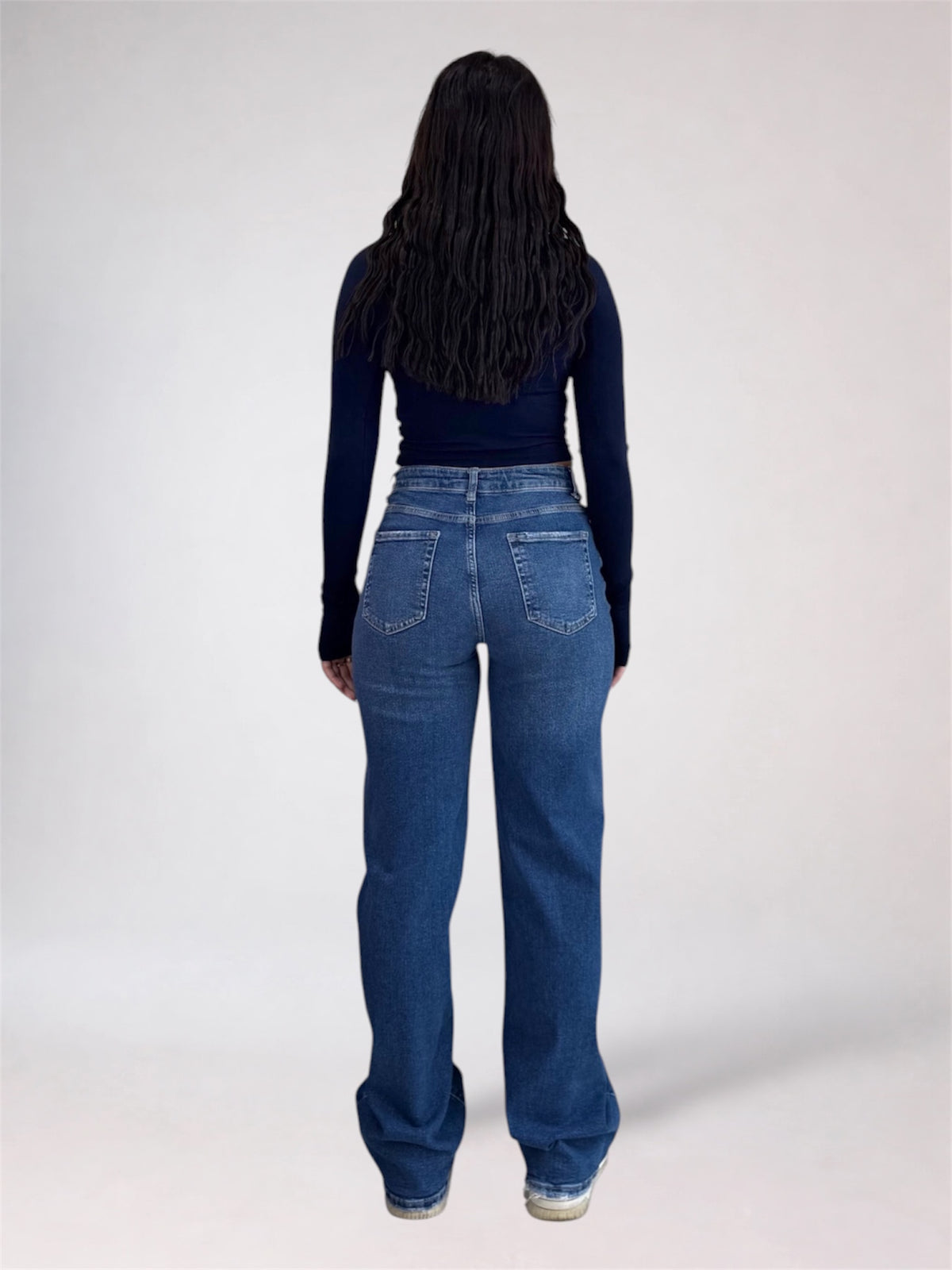 A person viewed from the back wearing a dark navy long-sleeve top and blue wide-leg jeans against a plain light gray studio background.