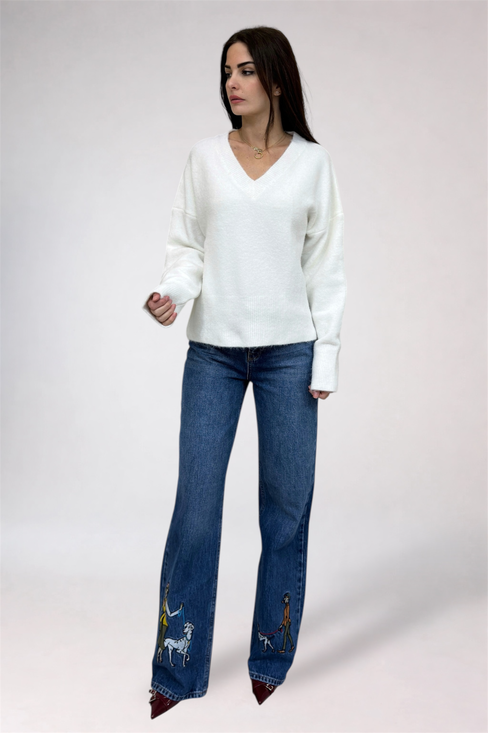 Woman wearing a white sweater and blue jeans with embroidery on a plain background