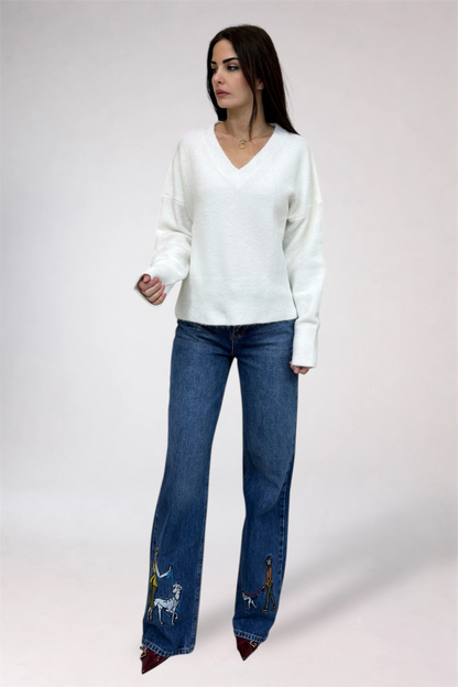 Woman wearing a white sweater and blue jeans with embroidery on a plain background