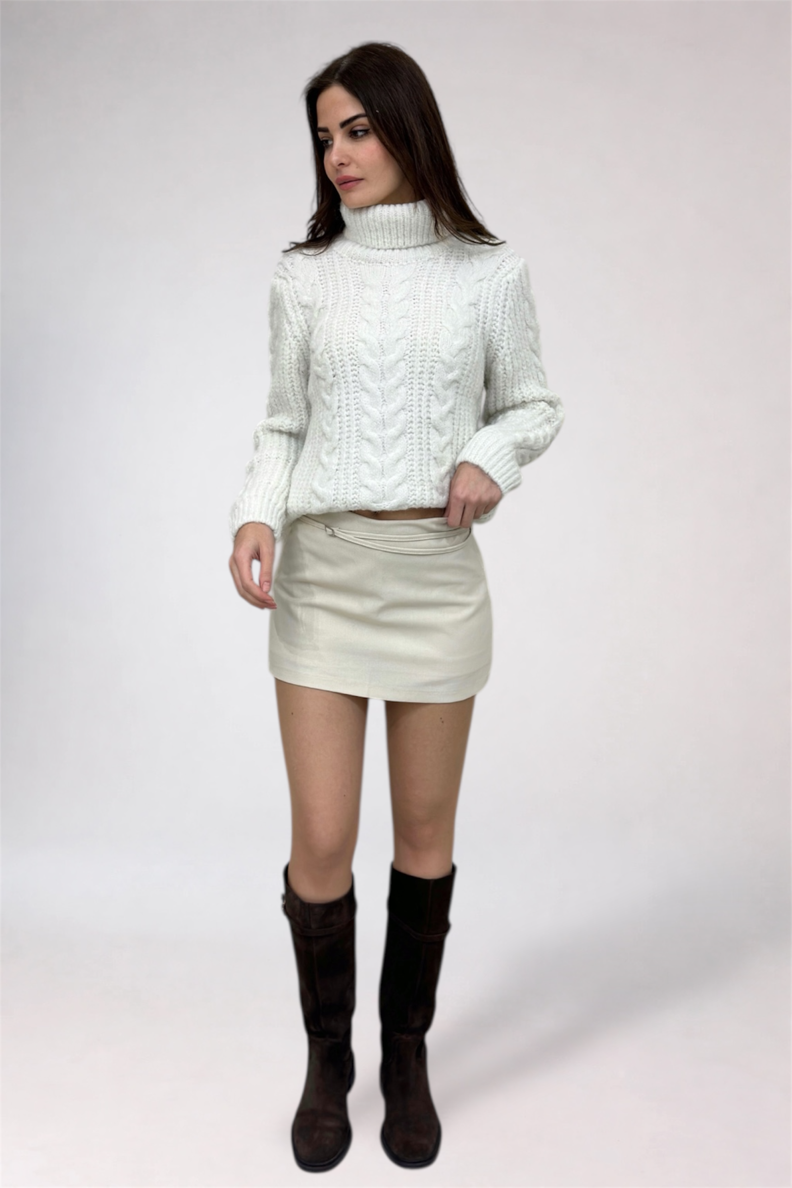 Woman wearing a white cable knit sweater, cream skirt, and black boots on a white background