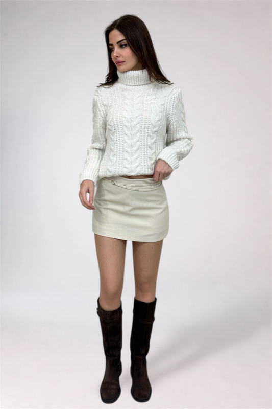 Woman wearing a white cable knit sweater, cream skirt, and black boots on a white background