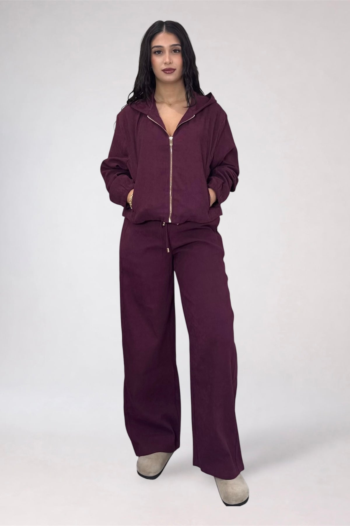 A woman modeling a matching deep plum purple tracksuit with a zip-up hoodie and wide-leg pants, standing against a light gray studio background.