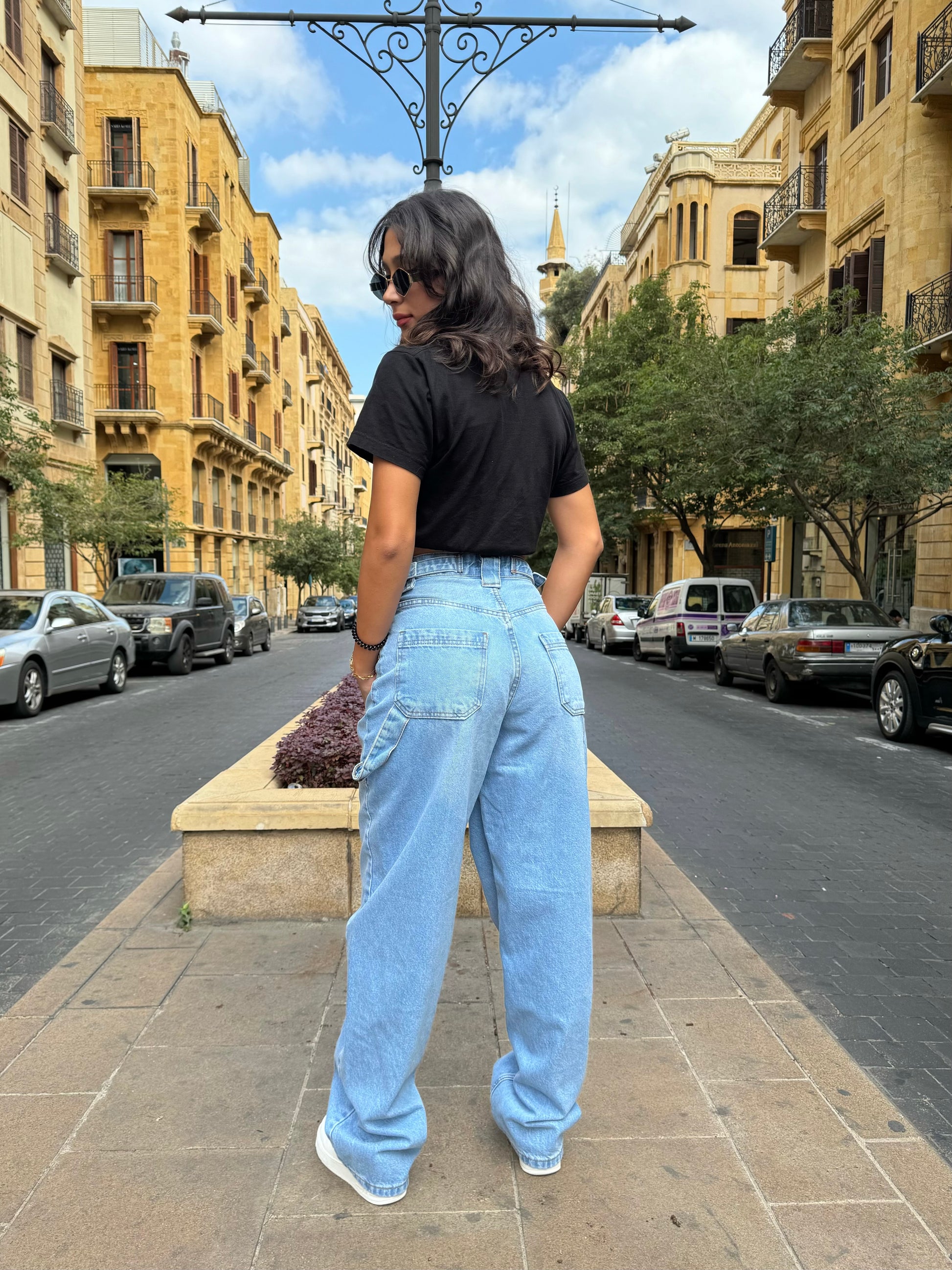Person wearing a black t-shirt and light blue jeans standing on a street with buildings and trees in the background.