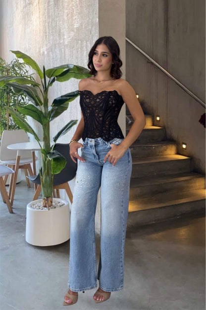 Woman in a black lace top and blue jeans standing in a modern indoor setting with plants and stairs.