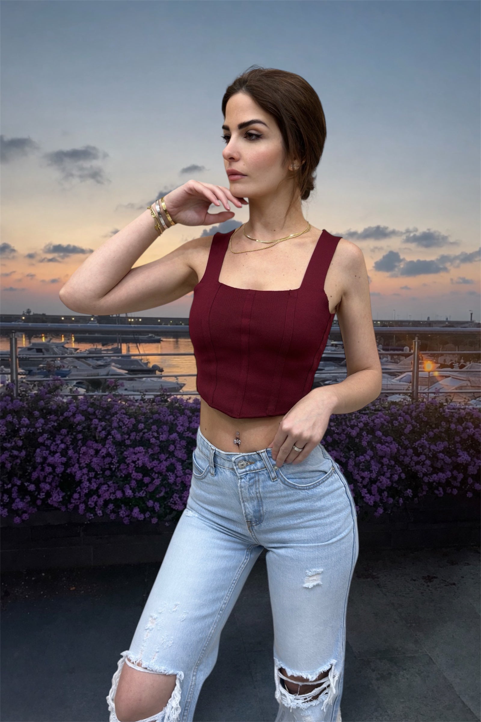 Woman in a burgundy top and light blue jeans standing outdoors with a scenic background.