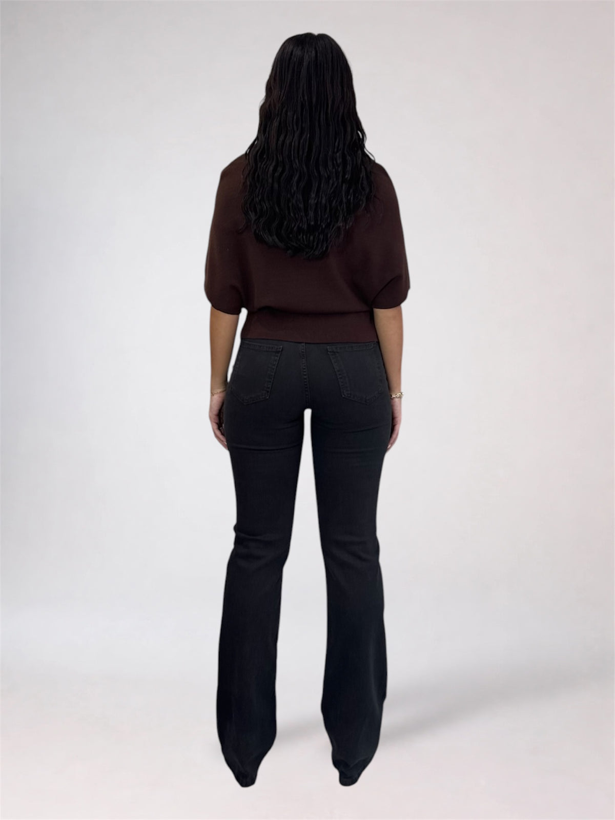 A woman shown from the back wearing a dark brown short-sleeve top and black flared jeans against a plain light gray studio background.