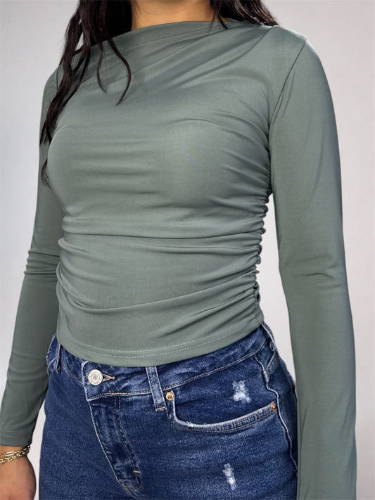 A woman wearing a fitted long-sleeve top in muted sage green with ruching at the waist, paired with blue jeans, against a plain light gray background.