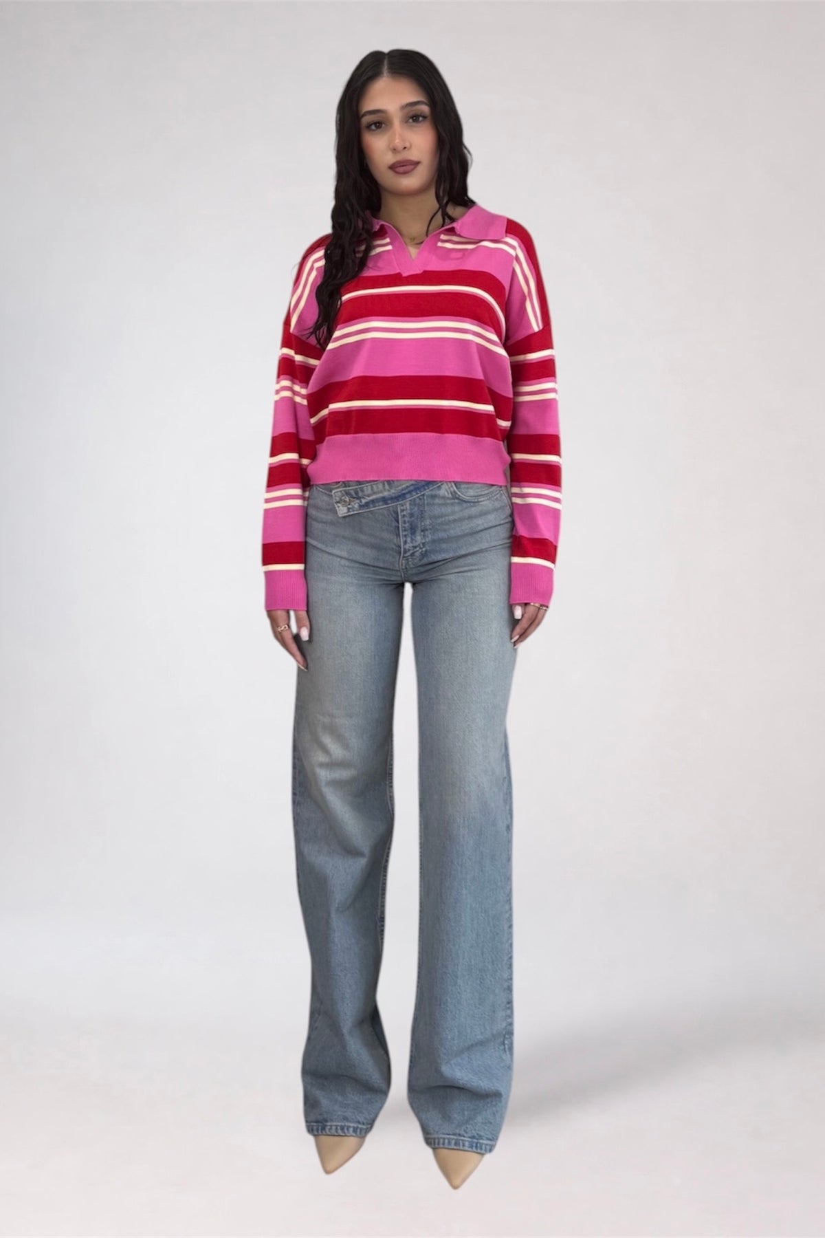 A model stands against a light gray studio background wearing a pink, red, and white striped long-sleeve knit polo sweater with light-wash blue jeans.
