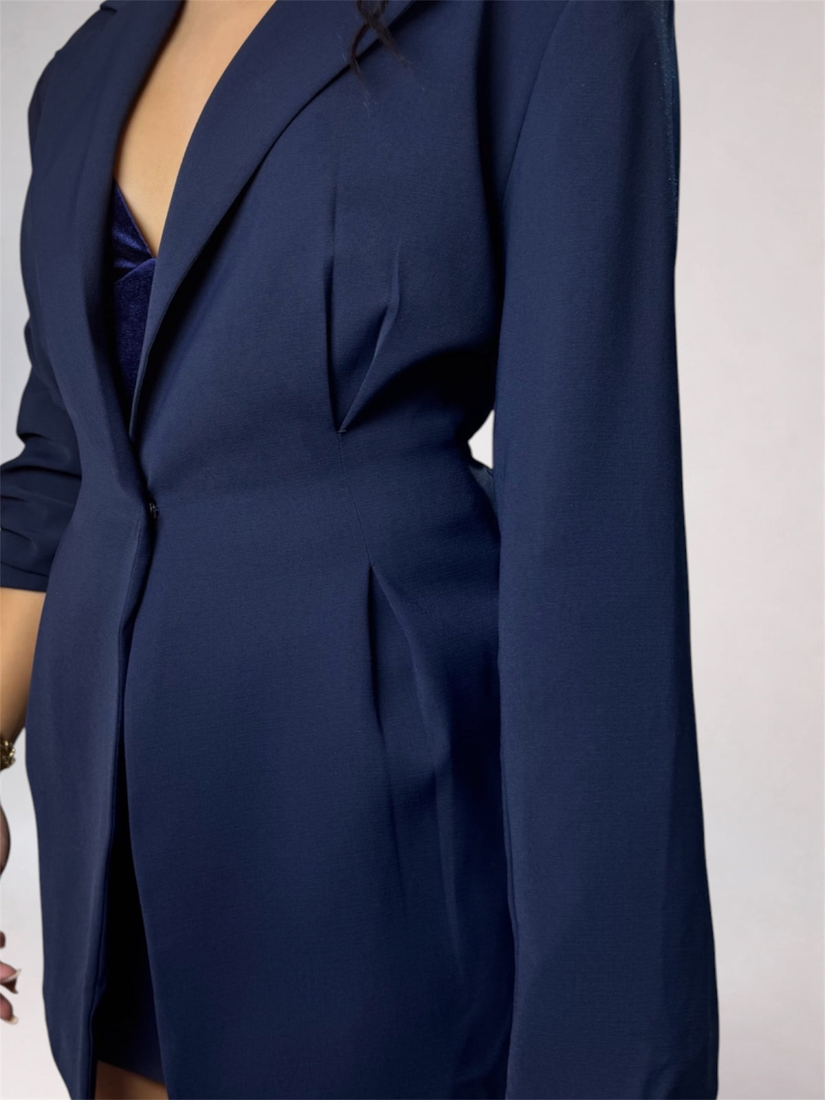 A close-up of a navy blue tailored blazer or dress with structured shoulders, a deep V-neckline, and front pleats, shown on a model against a light gray background.