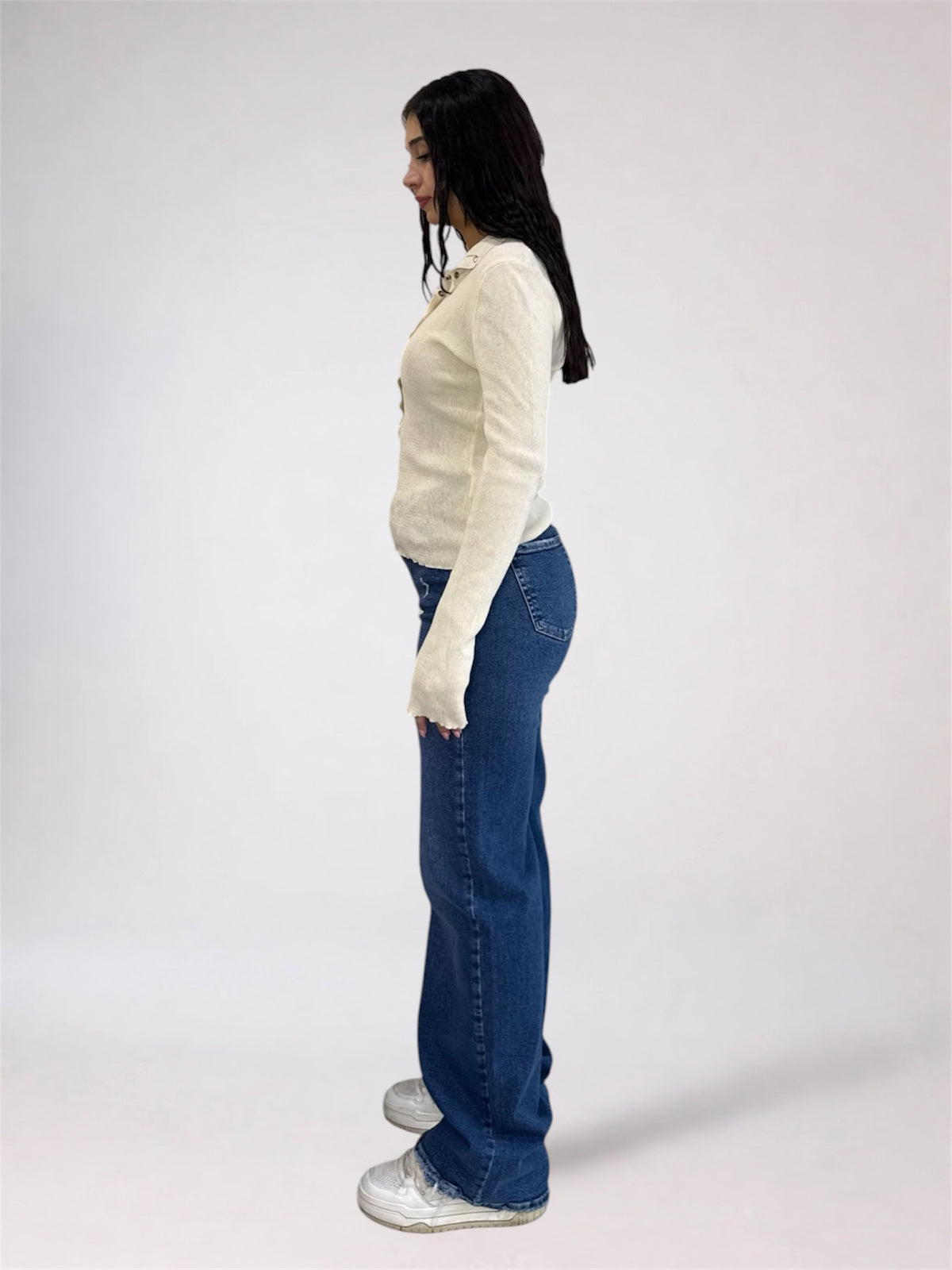 A woman standing in profile wearing a cream long-sleeve knit top, blue wide-leg jeans, and white sneakers against a plain light gray studio background.