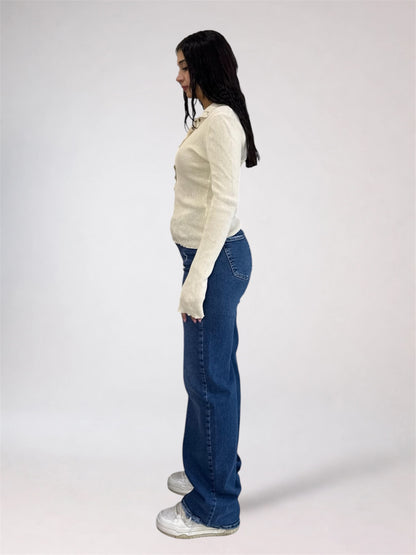 A woman standing in profile wearing a cream long-sleeve knit top, blue wide-leg jeans, and white sneakers against a plain light gray studio background.