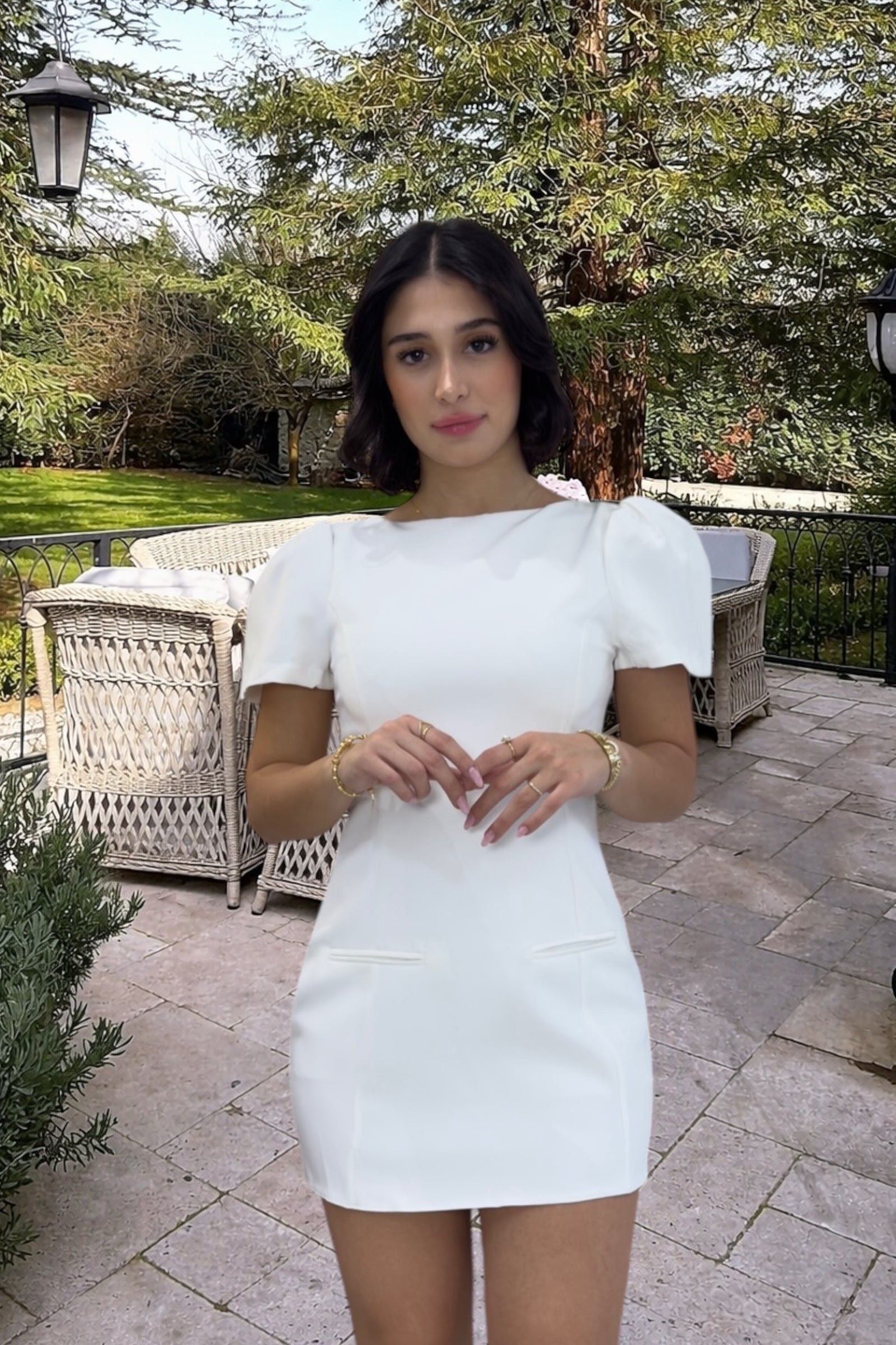 Woman in a white dress standing outdoors on a patio with greenery in the background