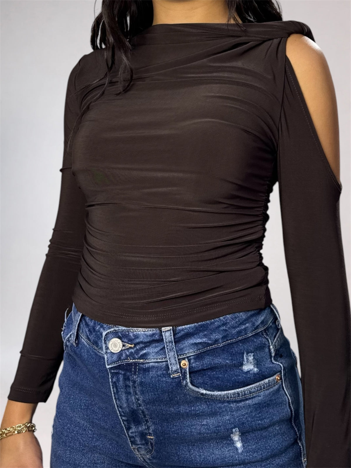 A woman wearing a dark brown long-sleeve top with a draped, ruched bodice and cold-shoulder cutouts, paired with blue jeans against a light gray background.