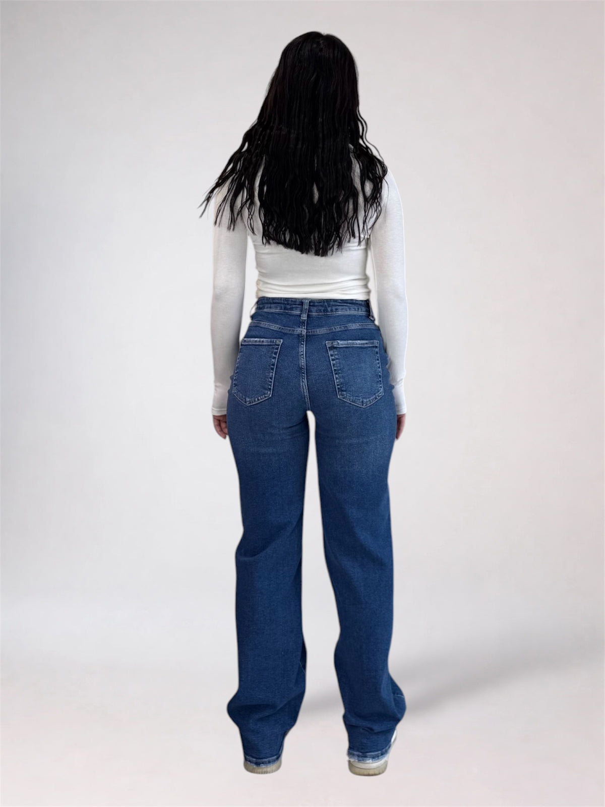 A model viewed from behind wearing high-waisted, wide-leg blue jeans and a white long-sleeve top, standing against a plain light gray studio background.