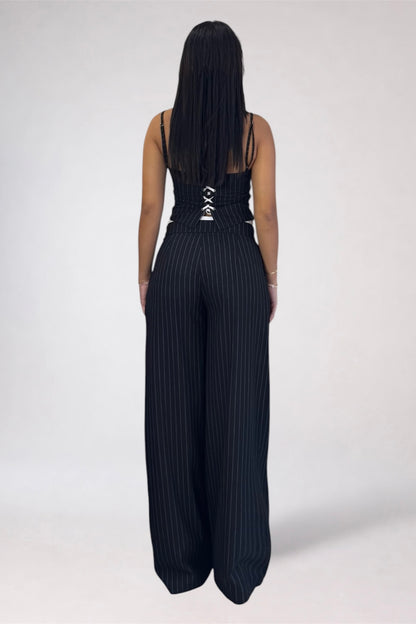 A woman viewed from behind wearing a black pinstriped jumpsuit with a lace-up back detail, set against a plain light gray studio background.