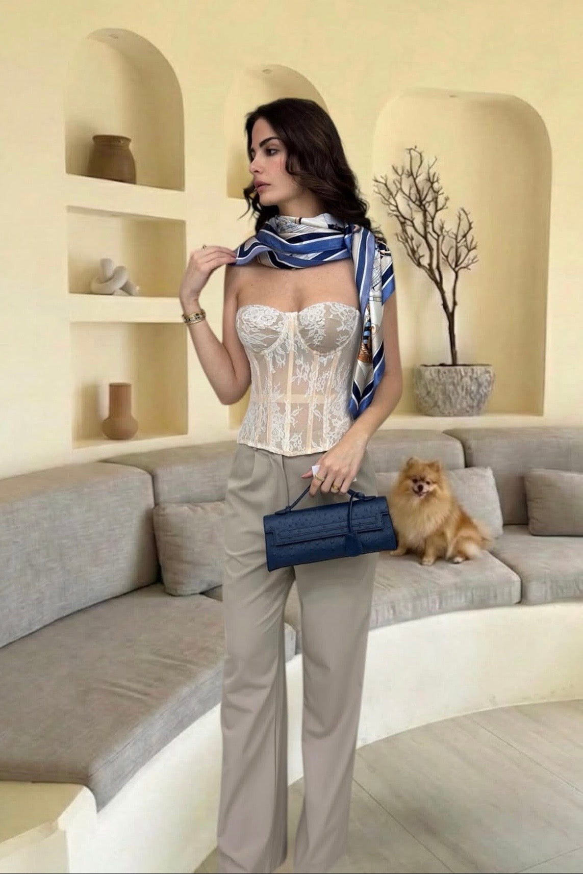 Woman in a stylish outfit with a scarf and handbag, standing in a modern living room.