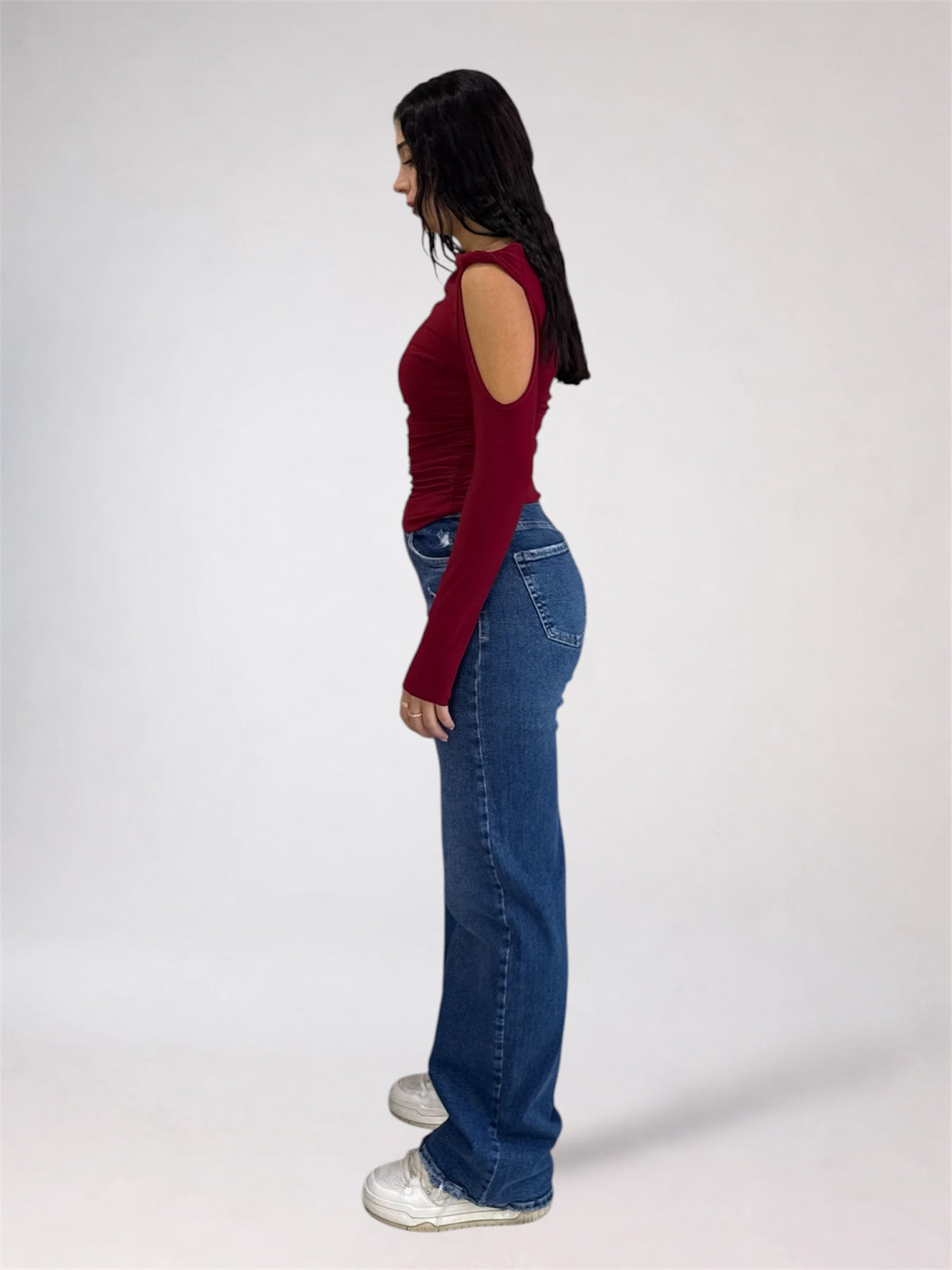 A woman shown in profile wearing a fitted long-sleeve cold-shoulder top in deep burgundy, blue jeans, and white sneakers, standing against a plain light gray studio background.