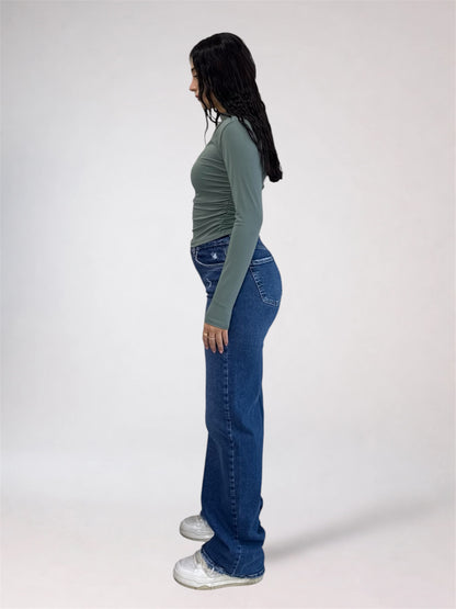 A woman shown in profile wearing a sage green long-sleeve top, blue wide-leg jeans, and white sneakers against a plain light gray studio background.