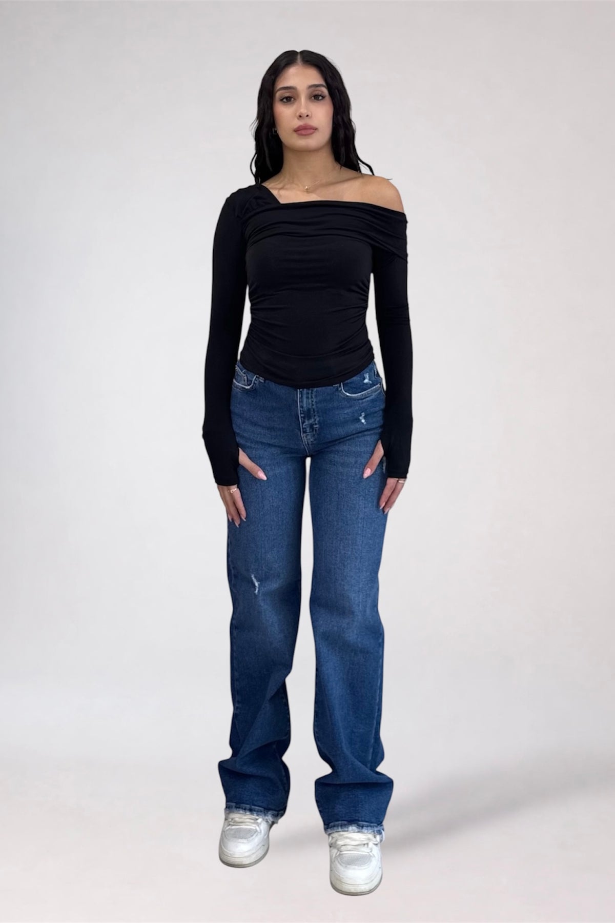 A woman modeling a black off-the-shoulder long-sleeve top paired with blue jeans and white sneakers, standing against a light gray studio background.