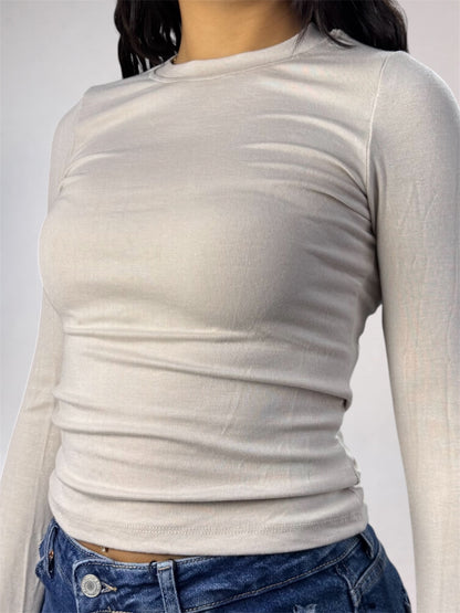 A close-up of a person wearing a fitted long-sleeve crew-neck top in a light warm gray or off-white tone, paired with blue jeans against a plain light gray background.