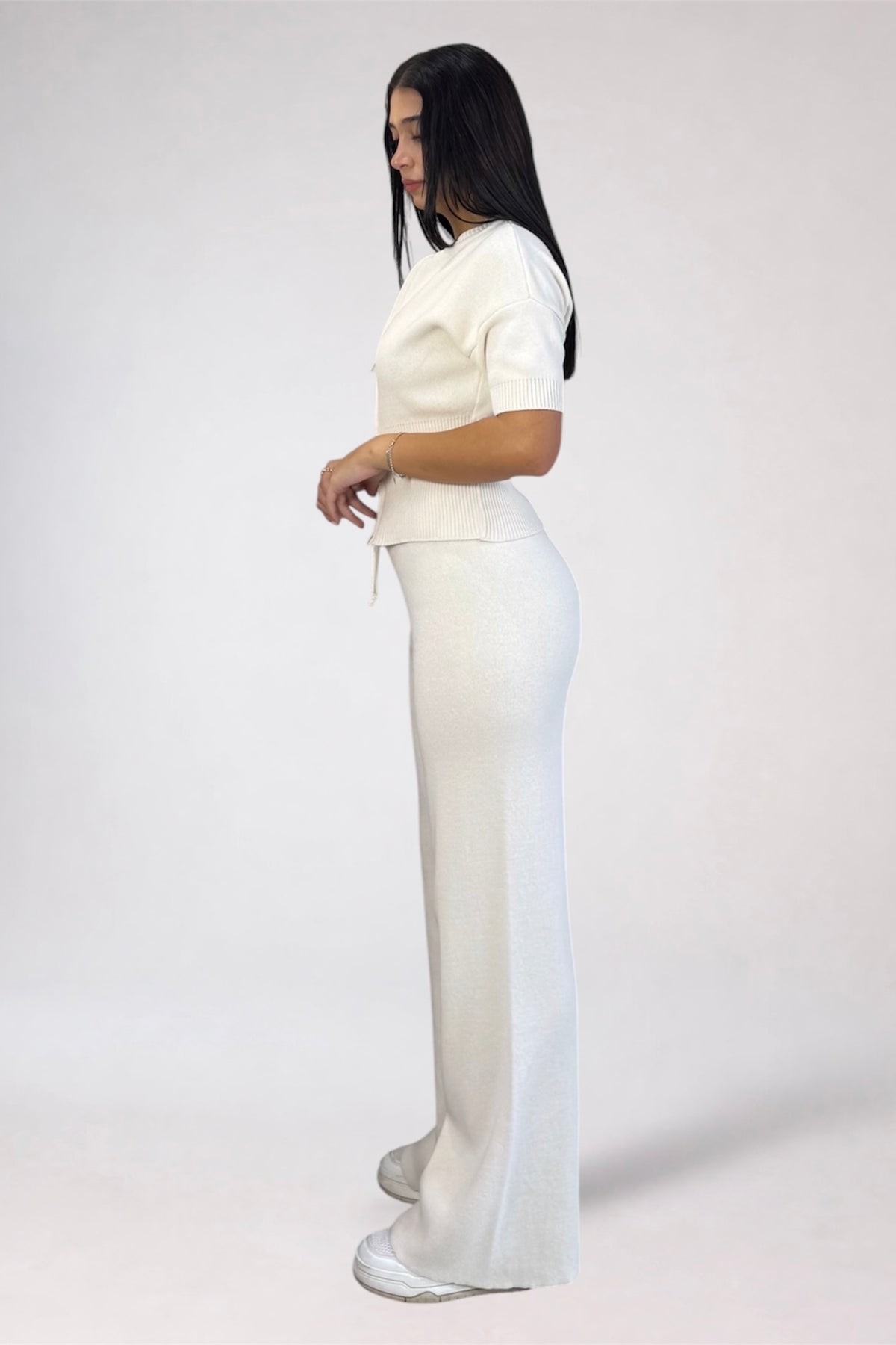 A woman modeling a matching white knit two-piece outfit consisting of a short-sleeve cropped top and high-waisted wide-leg pants, shown from a side profile against a light gray studio background.