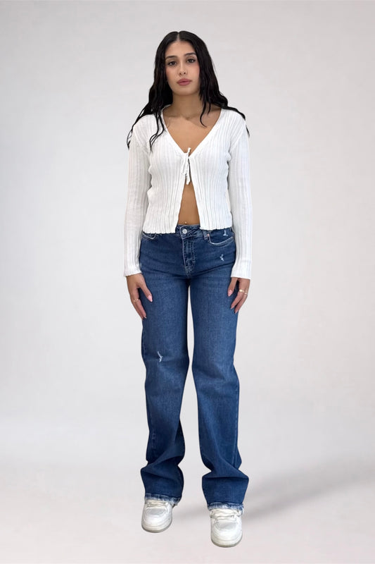 A model stands against a light gray studio background wearing a white ribbed long-sleeve tie-front cardigan, blue jeans, and white sneakers.