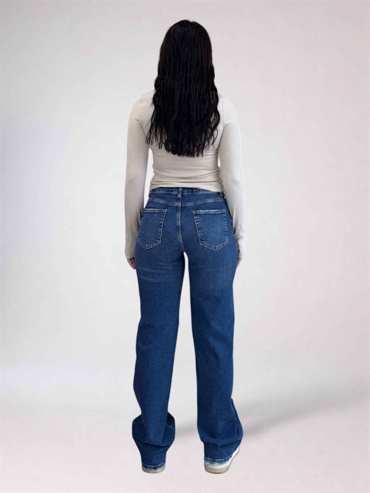 A person viewed from the back wearing a fitted light beige long-sleeve top and blue wide-leg jeans against a plain light gray studio background.