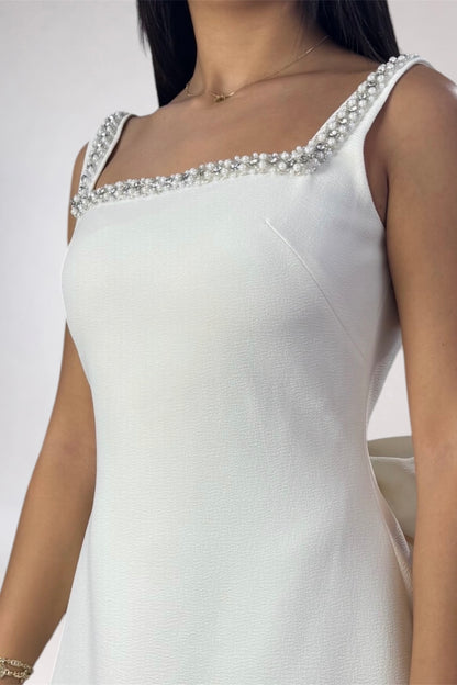 A close-up of a woman wearing a white sleeveless dress with a square neckline and a decorative border of pearls and clear embellishments along the neckline.